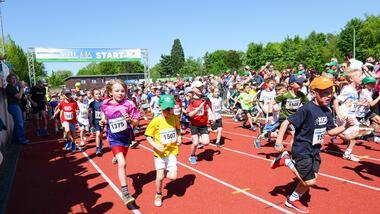 Start eines Kinderlaufes mit vielen laufenden Kindern auf einer roten Laufbahn, umgeben von Zuschauern und festlicher Atmosphäre.