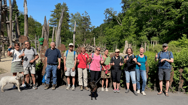 Gruppe von Wanderern mit Hunden posiert vor einer Holzskulptureninstallation im Freien an einem sonnigen Tag.
